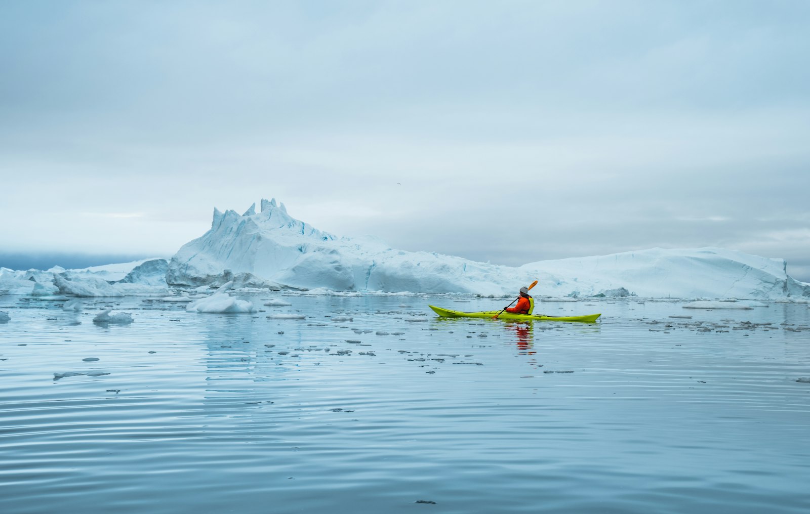 A person in a green sea kayak paddling among large icebergs in still water